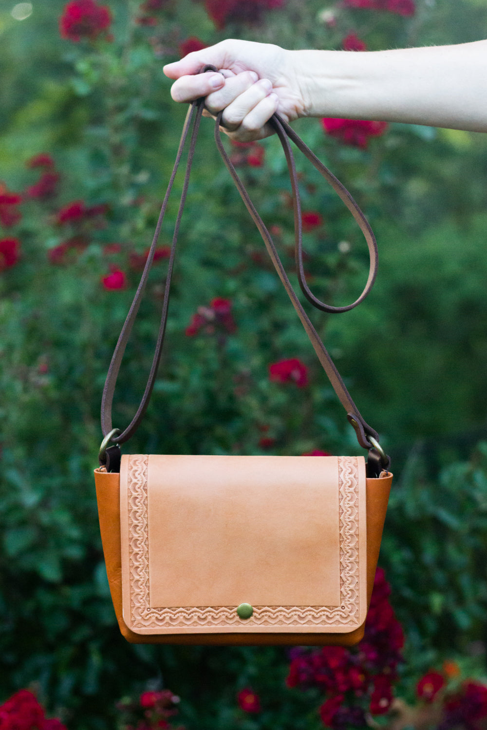 Tan leather handbag held by a hand with a blurred floral background
