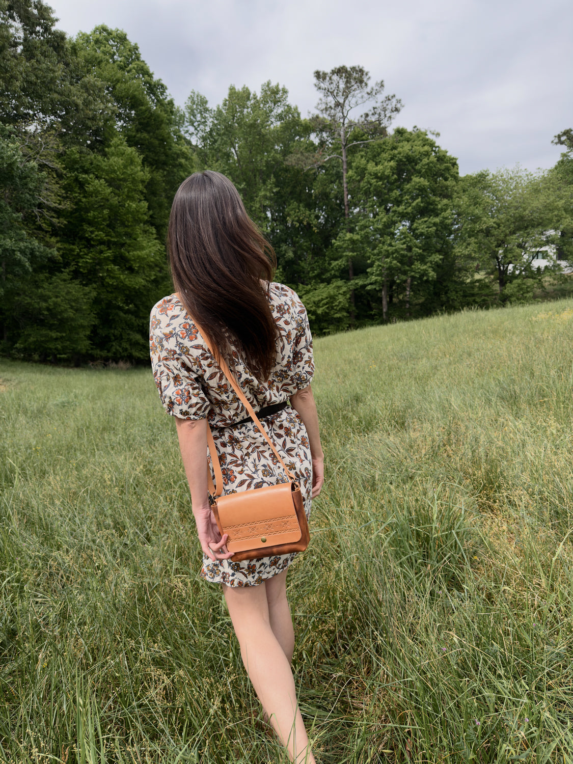 Woman walking through a grassy field holding a brown leather bag
