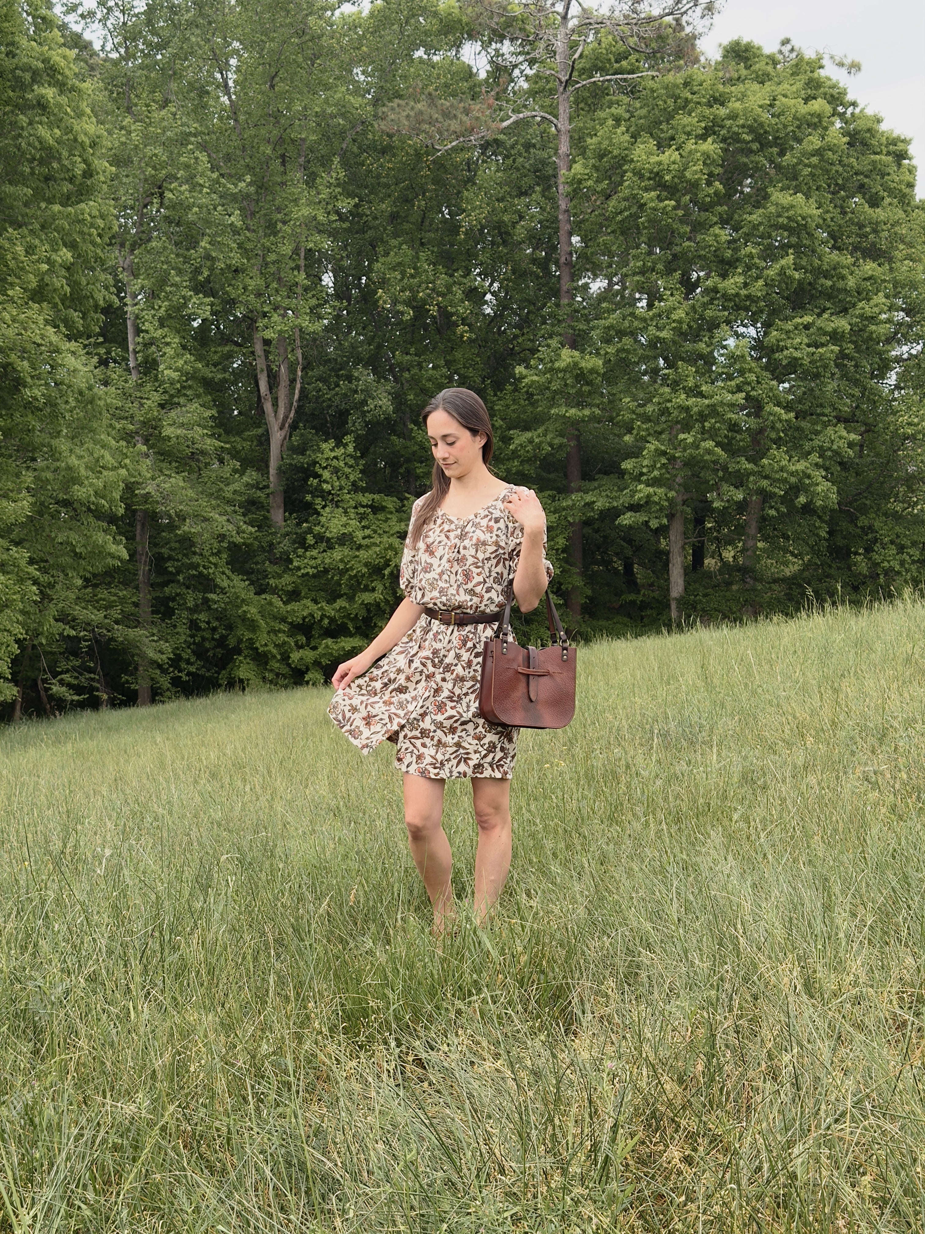 Woman in a floral dress and leather handbag standing in a grassy field with trees in the background